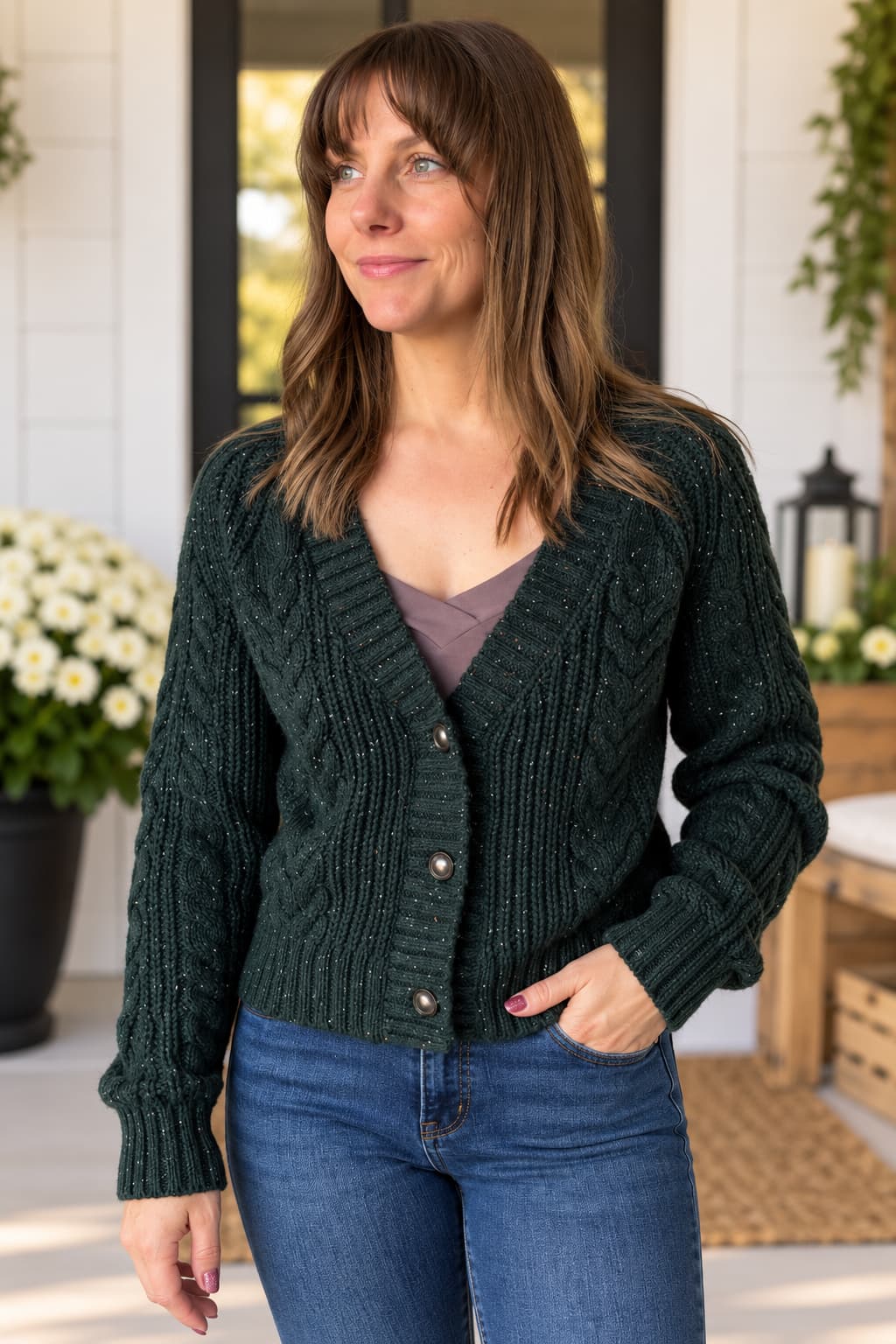 A woman wearing a dark forest green cable-knit cardigan sweater over a mauve top and blue jeans, standing in a bright, modern porch setting.