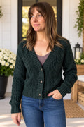 A woman wearing a dark forest green cable-knit cardigan sweater over a mauve top and blue jeans, standing in a bright, modern porch setting.
