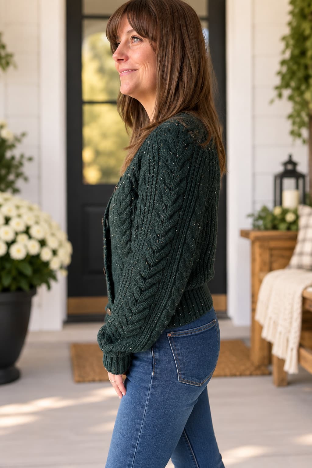 A woman wearing a dark forest green cable-knit cardigan sweater and blue jeans, standing on a sunny porch with white shiplap walls, potted flowers, and a wooden bench in the background.