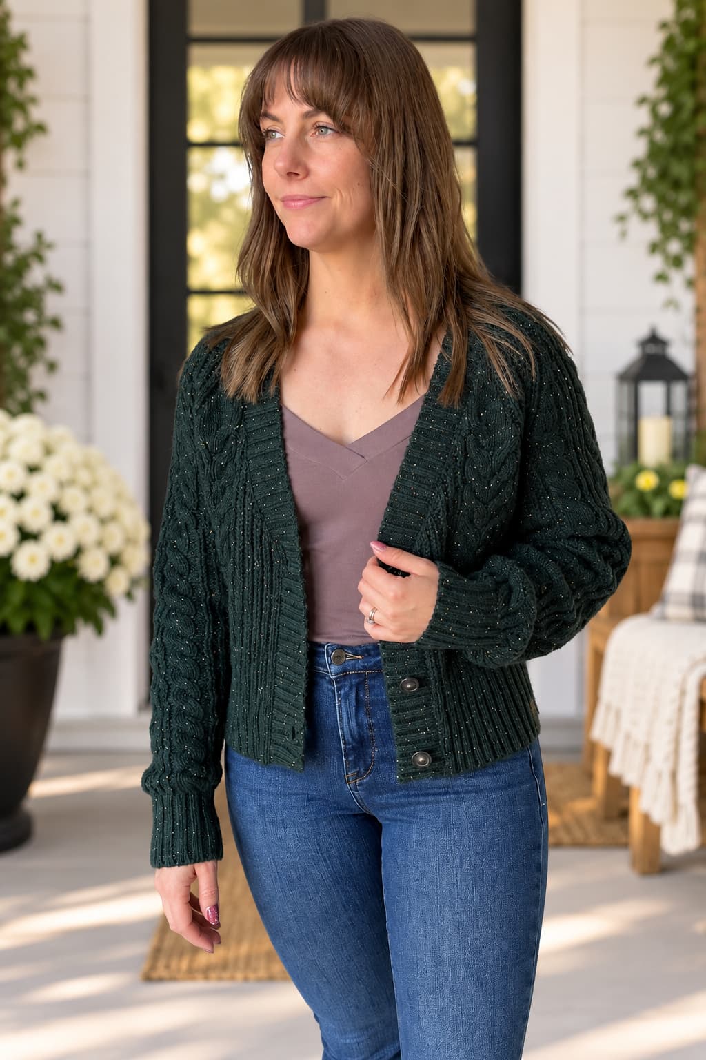 A woman wearing a dark green cable-knit cardigan over a mauve top and blue jeans, standing on a porch with potted plants and a lantern in the background.