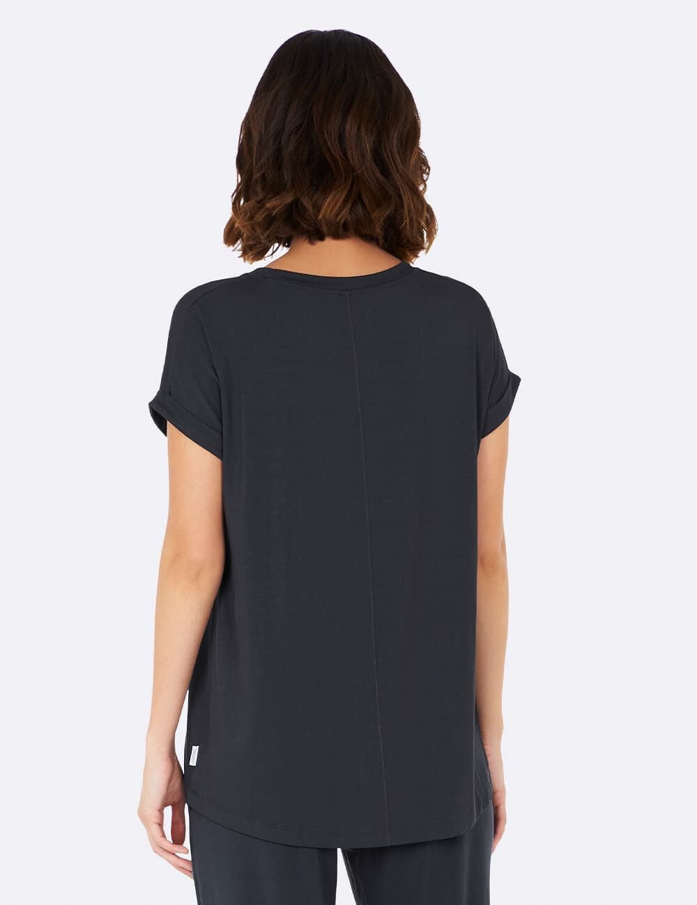A woman viewed from the back wearing a dark blue gray short-sleeve top with a relaxed fit and a small white tag near the lower left hem, set against a plain light gray background.