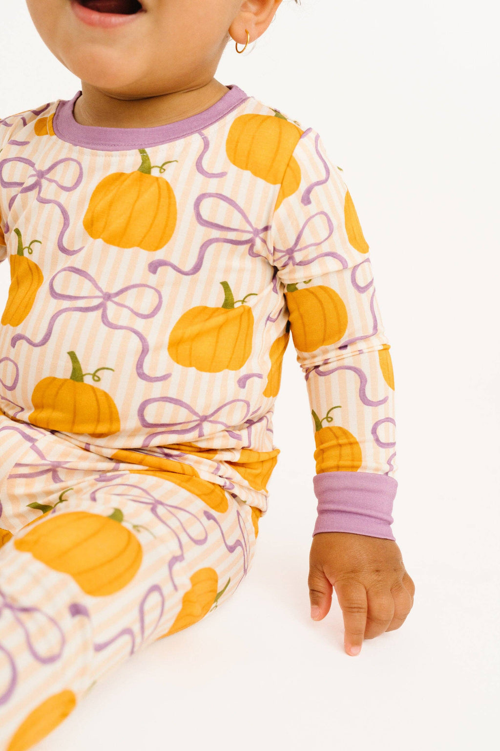 A close-up of a baby wearing a long-sleeve pajama top and matching pants with an orange pumpkin and purple ribbon pattern on a cream striped background, set against a plain white studio background.