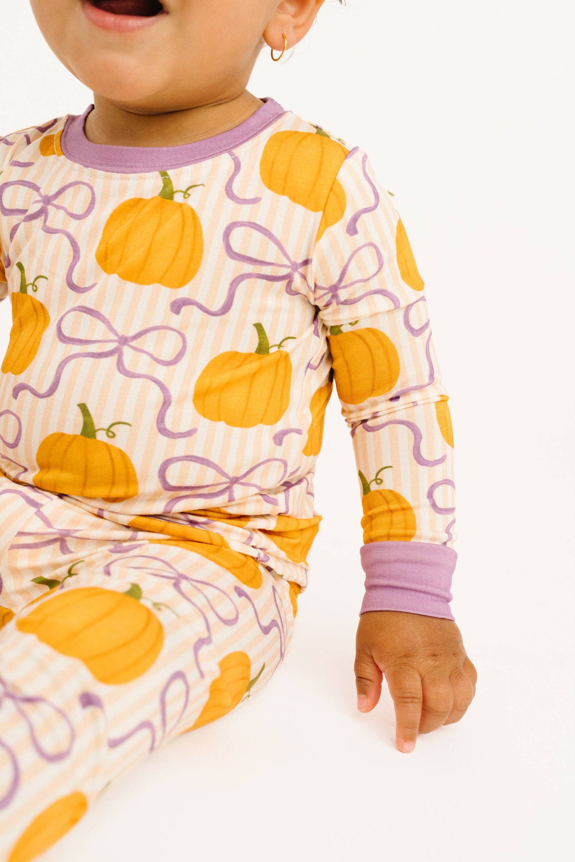 A close-up of a baby wearing a long-sleeve pajama top and matching pants with an orange pumpkin and purple ribbon pattern on a cream striped background, set against a plain white studio background.