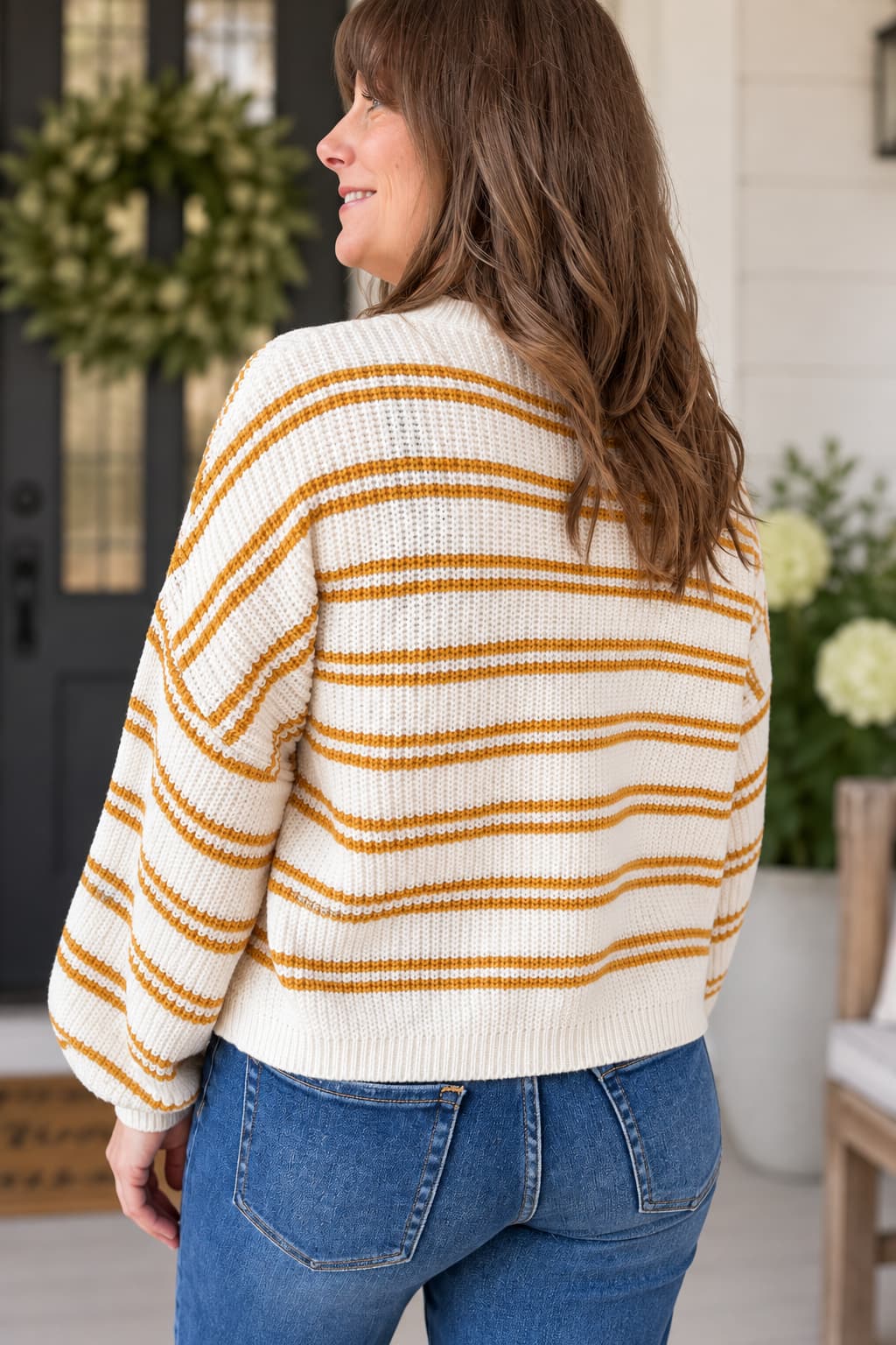 A woman wearing a cream knit sweater with mustard yellow horizontal stripes, shown from the back as she stands on a porch wearing blue jeans.