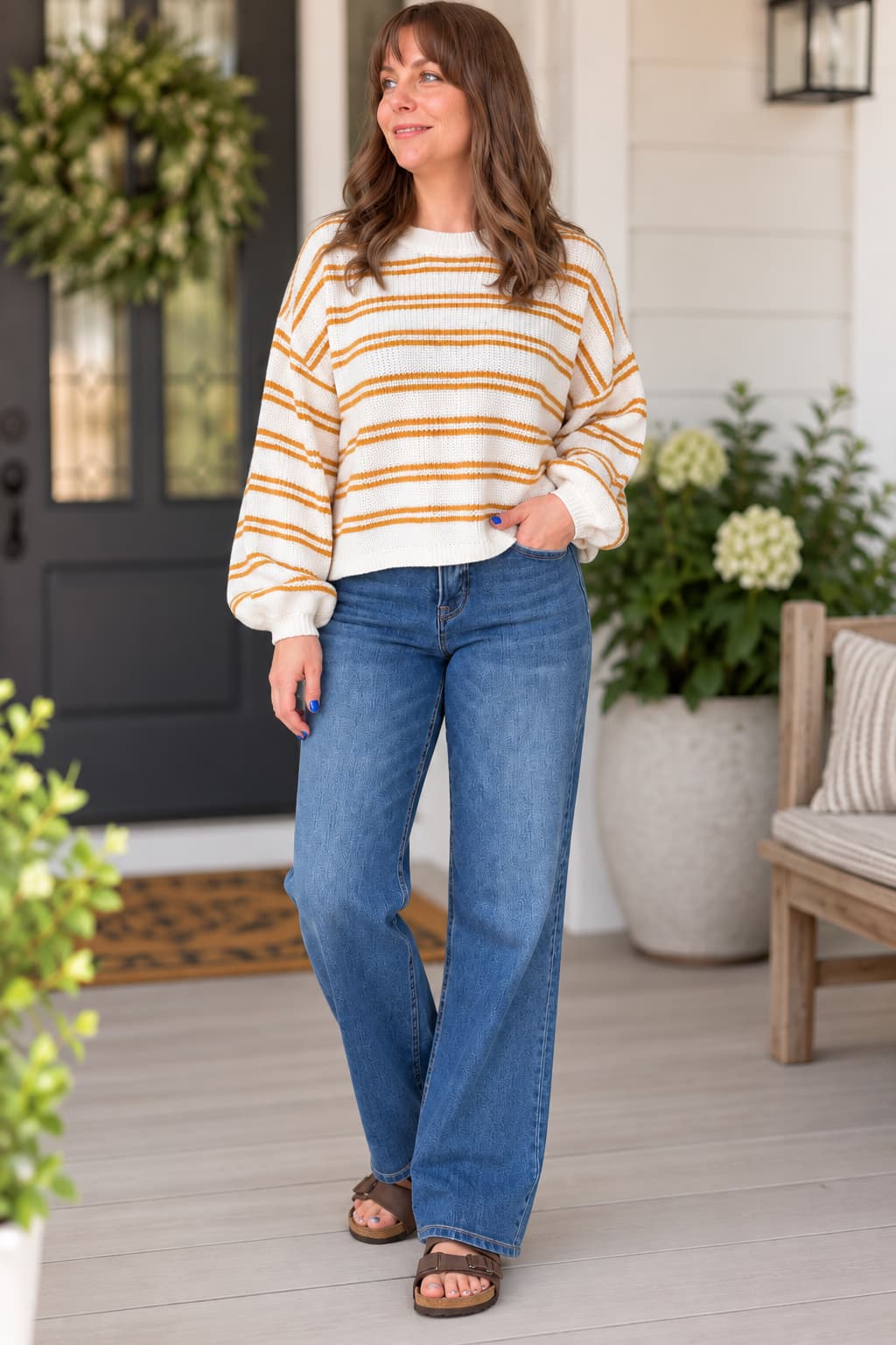 A woman stands on a porch wearing a cream and mustard yellow striped long-sleeve knit sweater, blue jeans, and brown sandals.
