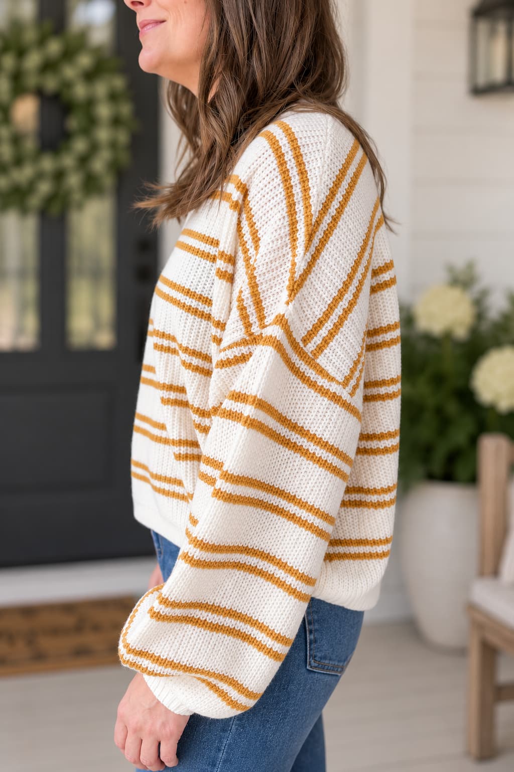 A woman wearing a chunky knit sweater with horizontal mustard yellow stripes on an off-white background, paired with blue jeans, standing in a bright, modern porch setting.