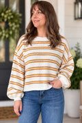 A woman wearing a cream-colored knit sweater with horizontal mustard yellow stripes, paired with blue jeans, standing in front of a softly blurred porch background.
