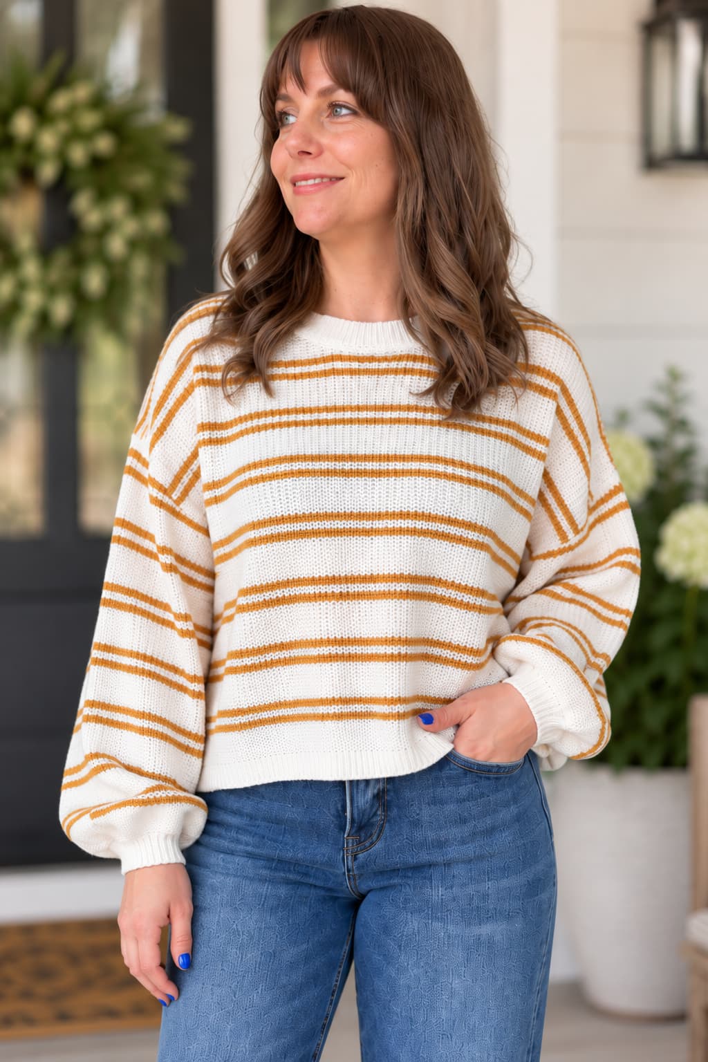 A woman wearing a cream-colored knit sweater with horizontal mustard yellow stripes, paired with blue jeans, standing in front of a softly blurred porch background.