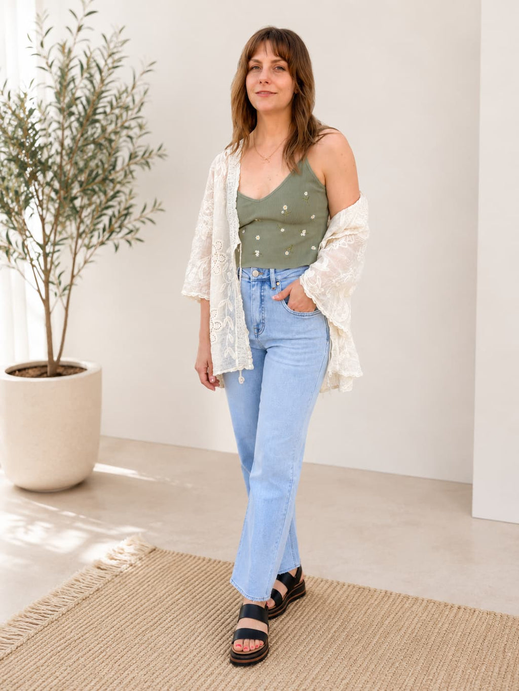 Woman standing in a room wearing sage daisy embroidered tank and kimono with blue denim