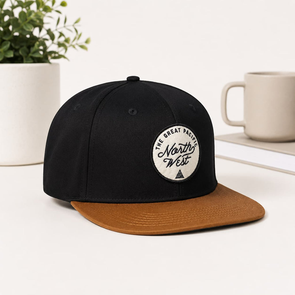 A black flat-brim cap with a brown visor and a circular white patch reading 'The Great Pacific North West' on the front, styled on a white surface with a plant and mug in the background.