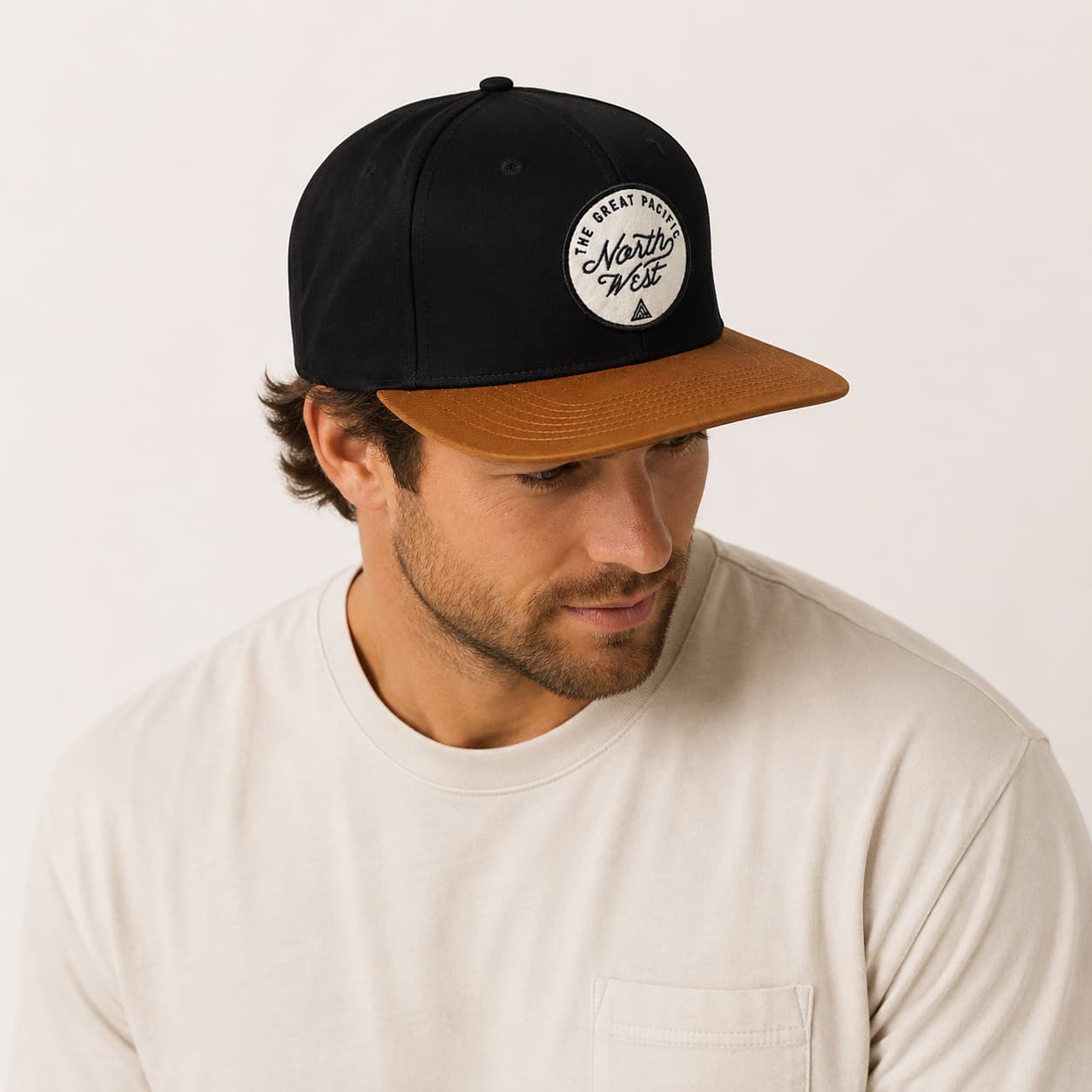A man wearing a black and brown flat-brim snapback cap with a circular 'The Best of North West' logo patch on the front, paired with a cream-colored t-shirt against a plain off-white background.