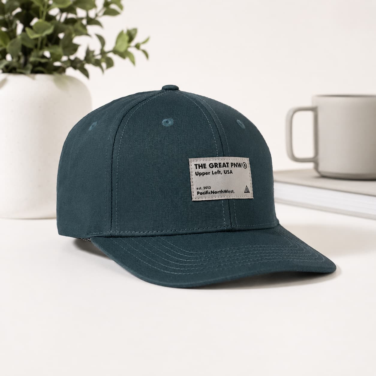 A dark teal baseball cap with a white rectangular patch reading 'THE GREAT PINNY Upper Left, USA' and 'PaisleyHerbBitty' at the bottom, displayed on a white surface with a mug and plant in the background.