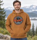 A man wearing a tan pullover hoodie with a circular 'Rainier Mountain Fresh' graphic on the chest, standing outdoors in front of a scenic mountain and lake background.