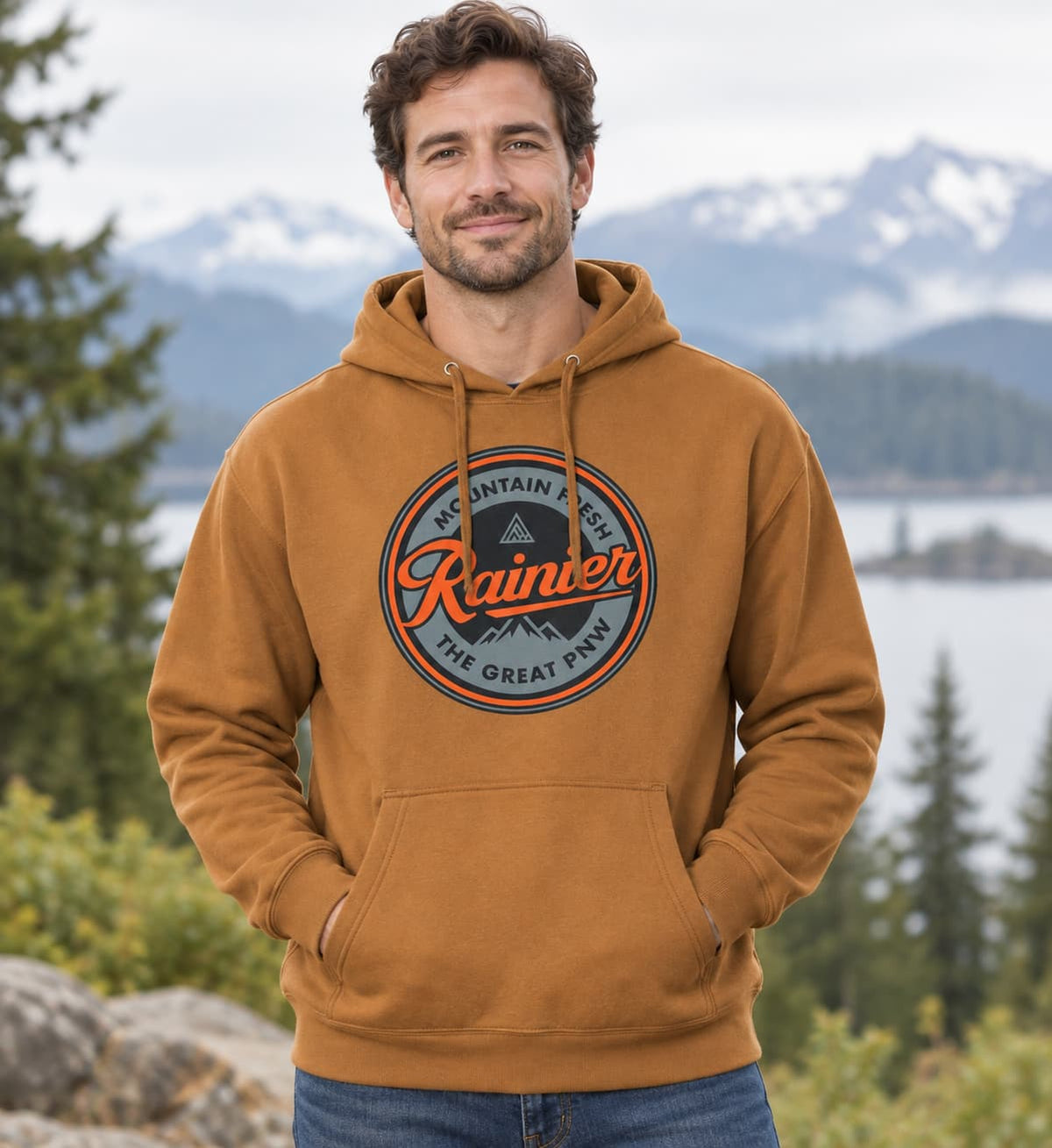 A man wearing a tan pullover hoodie with a circular 'Rainier Mountain Fresh' graphic on the chest, standing outdoors in front of a scenic mountain and lake background.