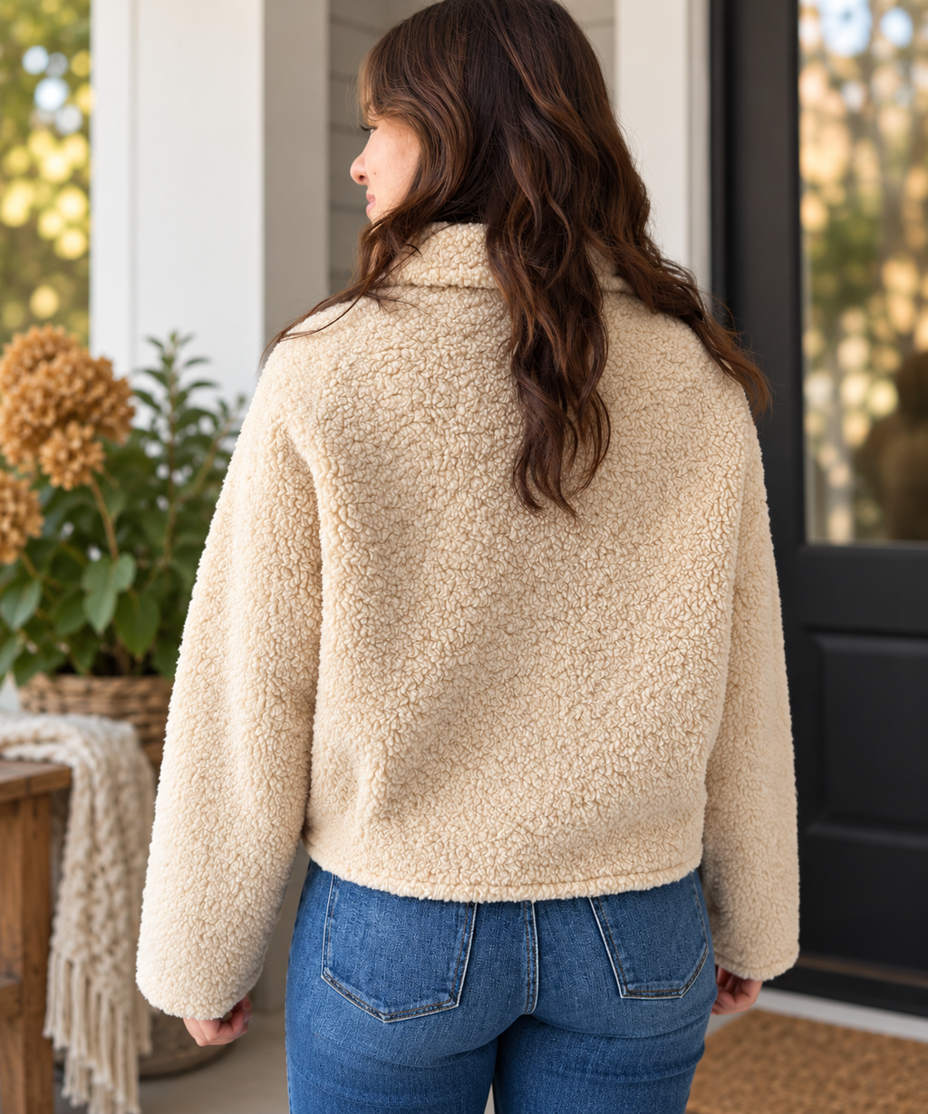 A woman viewed from the back wearing a cream-colored sherpa fleece jacket and blue jeans, standing on a porch.