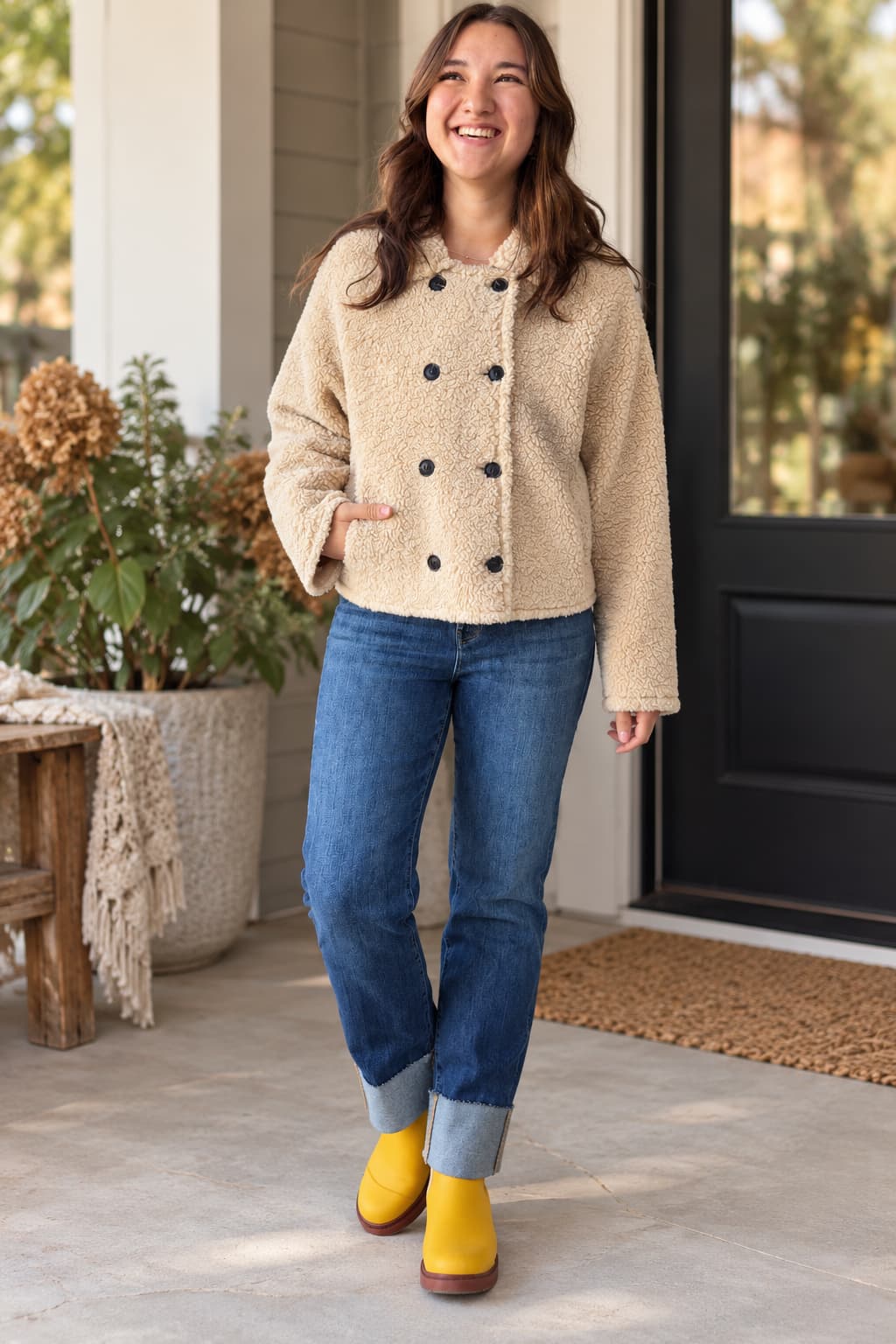 A woman stands on a porch wearing a beige sherpa-style cropped jacket with black buttons, blue jeans, and bright yellow rain boots.