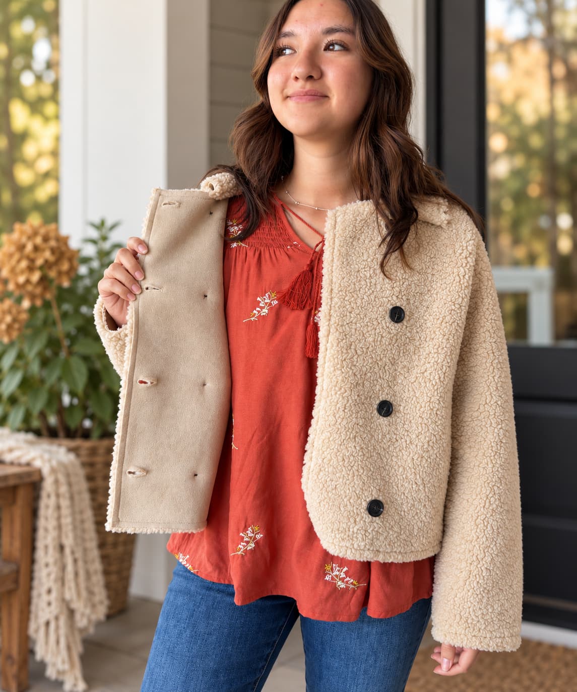 A woman wearing a beige sherpa fleece button-front jacket over a rust-orange embroidered top and blue jeans, standing on a porch.