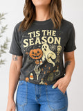 A woman wearing a dark charcoal gray graphic t-shirt with a Halloween theme and the text 'TIS THE SEASON' in cream. The graphic features a jack-o'-lantern, ghost, skeleton, and candy corn.