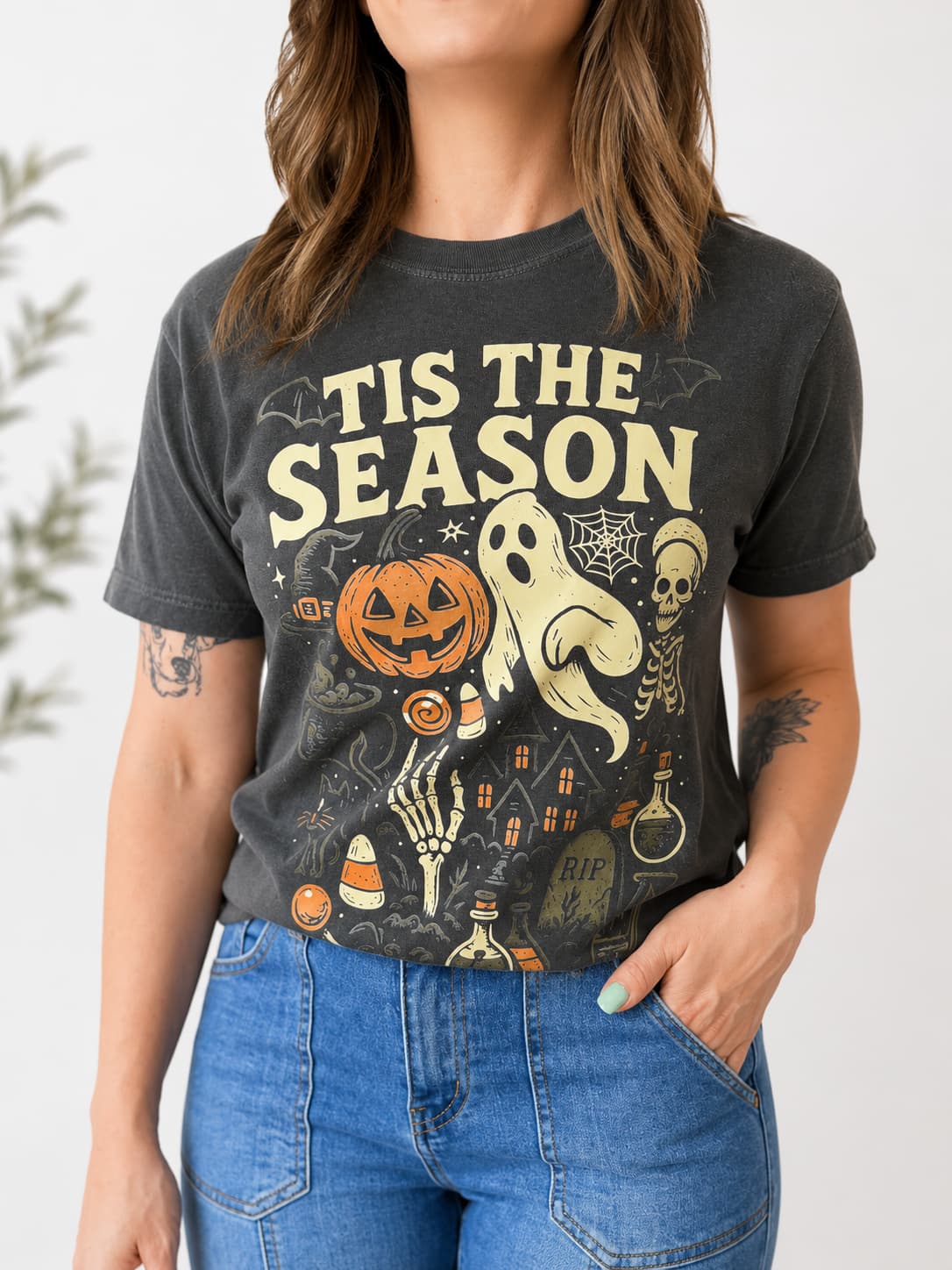 A woman wearing a dark charcoal gray graphic t-shirt with a Halloween theme and the text 'TIS THE SEASON' in cream. The graphic features a jack-o'-lantern, ghost, skeleton, and candy corn.