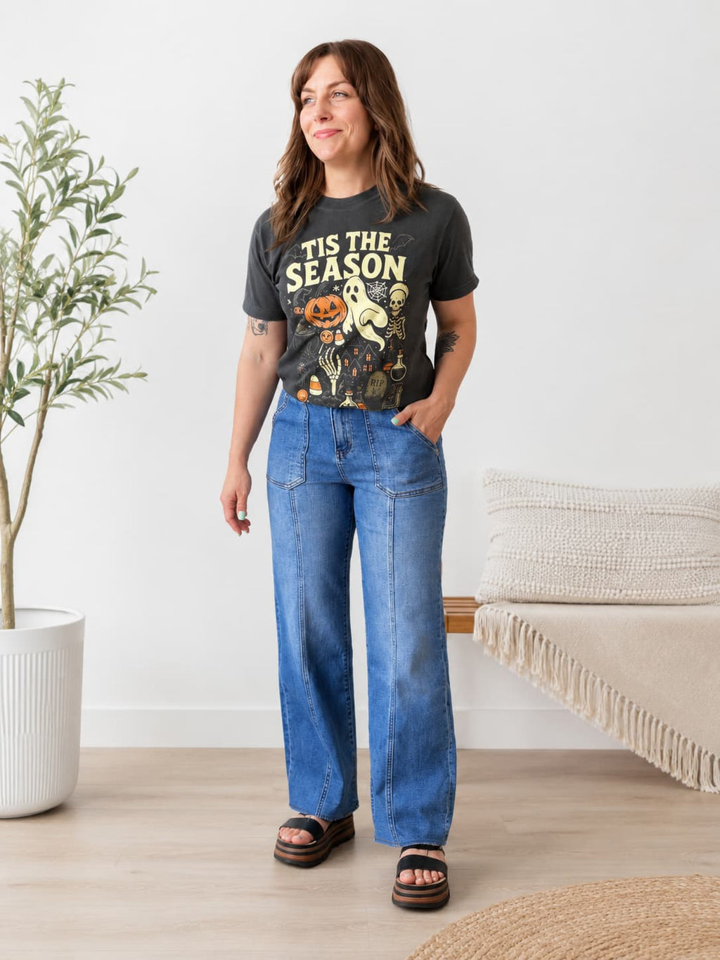 A woman modeling a dark charcoal graphic t-shirt with 'TIS THE SEASON' and a Halloween illustration, paired with wide-leg blue jeans and black slide sandals in a bright, modern room.