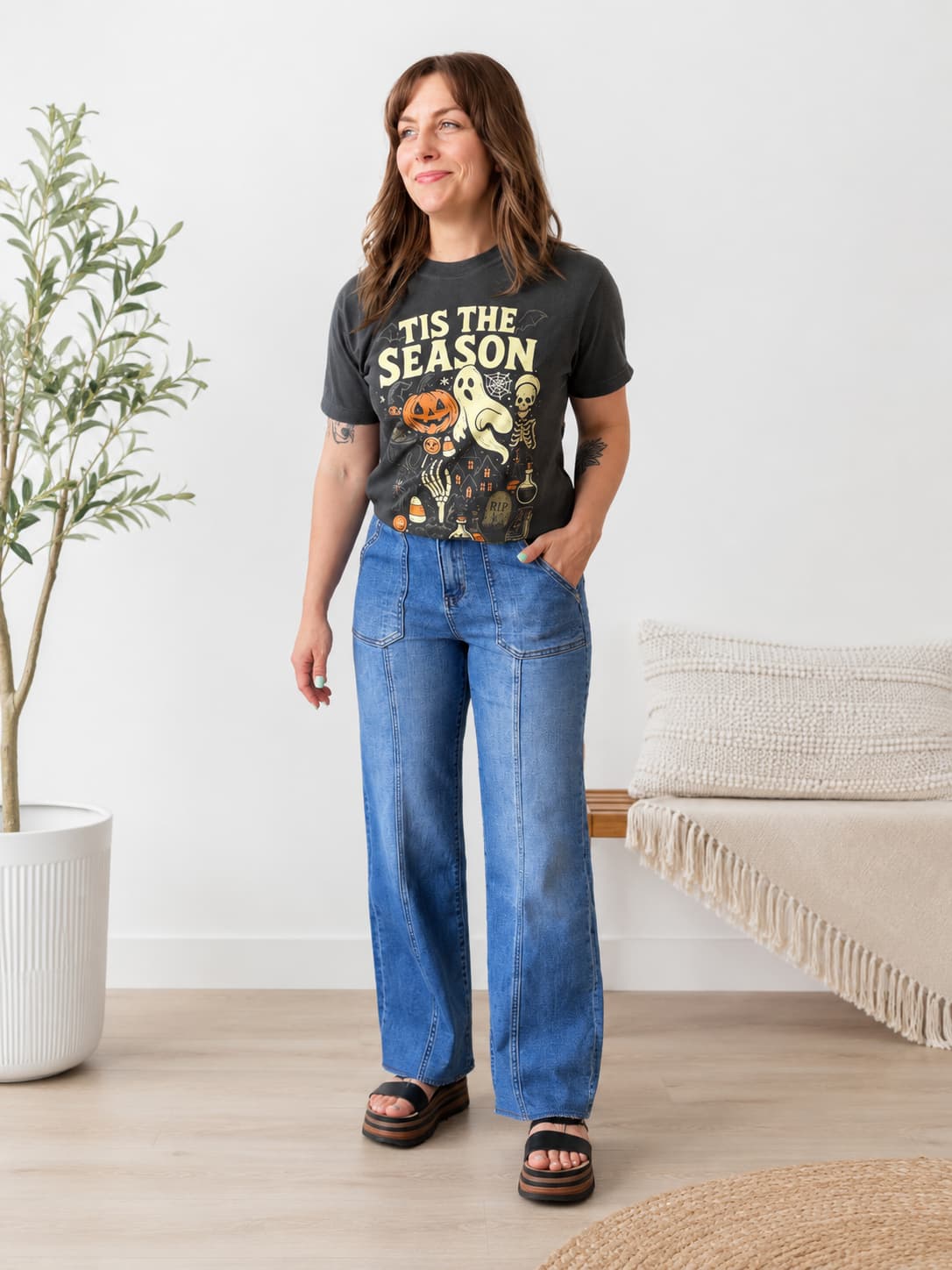 A woman modeling a dark charcoal graphic t-shirt with 'TIS THE SEASON' and a Halloween illustration, paired with wide-leg blue jeans and black slide sandals in a bright, modern room.