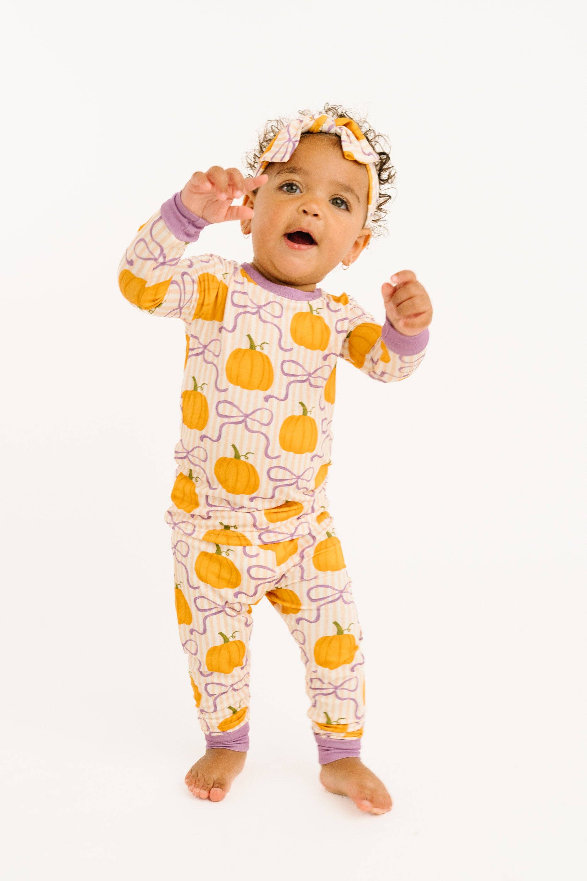A baby wearing a long-sleeve pajama set with an orange pumpkin and purple bow pattern on a cream background, along with a matching headband, standing against a white studio background.