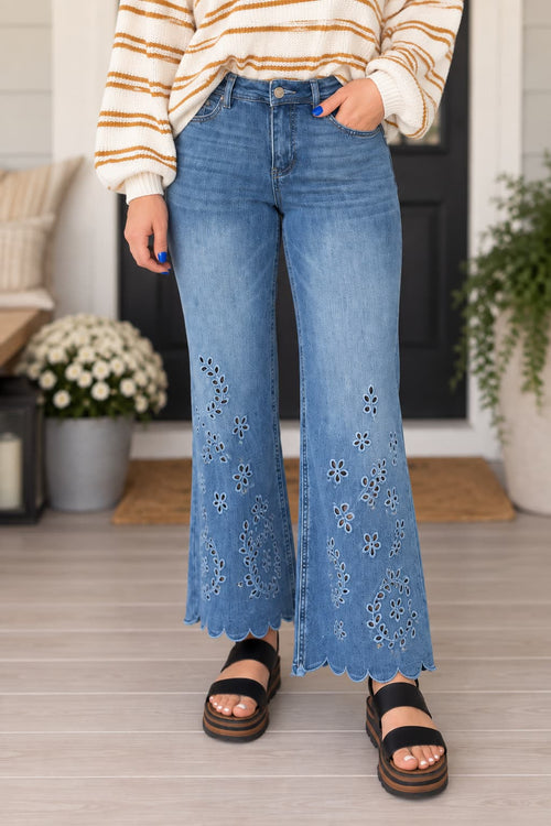 A person wearing light blue wide-leg jeans with floral eyelet embroidery down the front, paired with a cream and tan striped sweater and black platform sandals, standing in a home entryway.