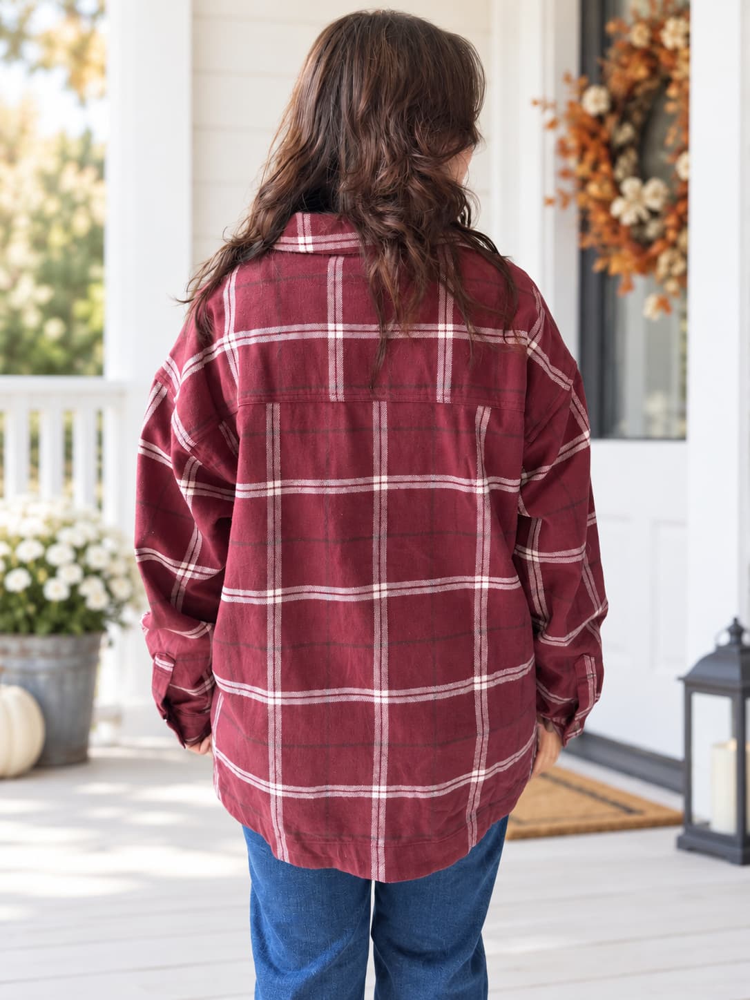 A woman viewed from behind wearing an oversized burgundy plaid flannel shirt and blue jeans, standing on a white porch with fall decorations in the background.