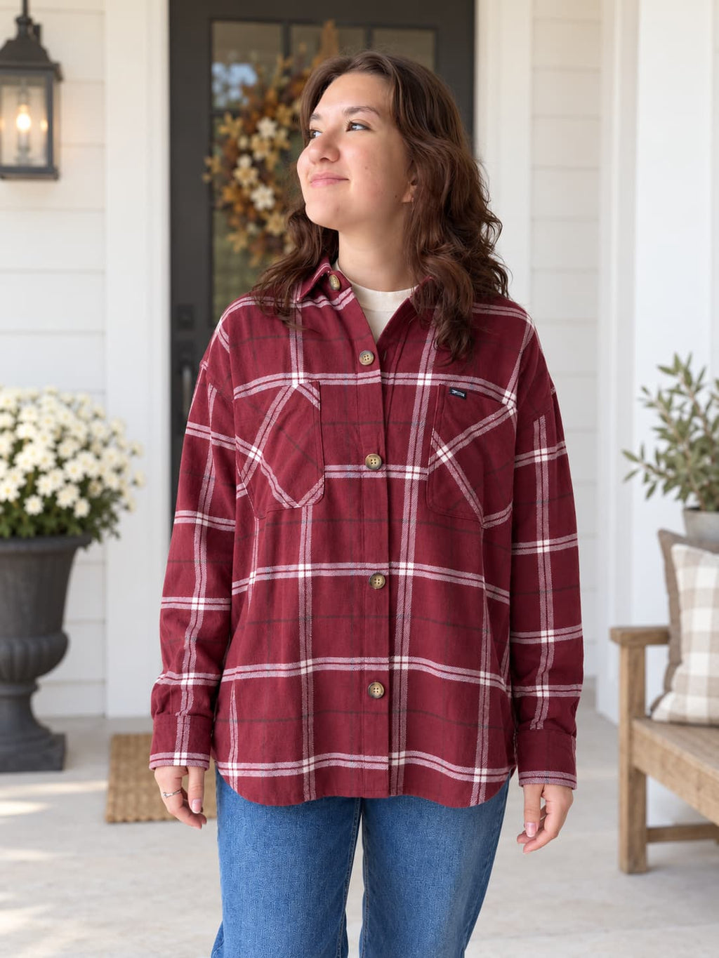 A woman wearing a burgundy and white plaid button-up flannel shirt with blue jeans, standing on a white porch with potted flowers and a wooden bench in the background.