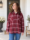 A woman wearing a burgundy and white plaid button-up flannel shirt with blue jeans, standing on a white porch with potted flowers and a wooden bench in the background.