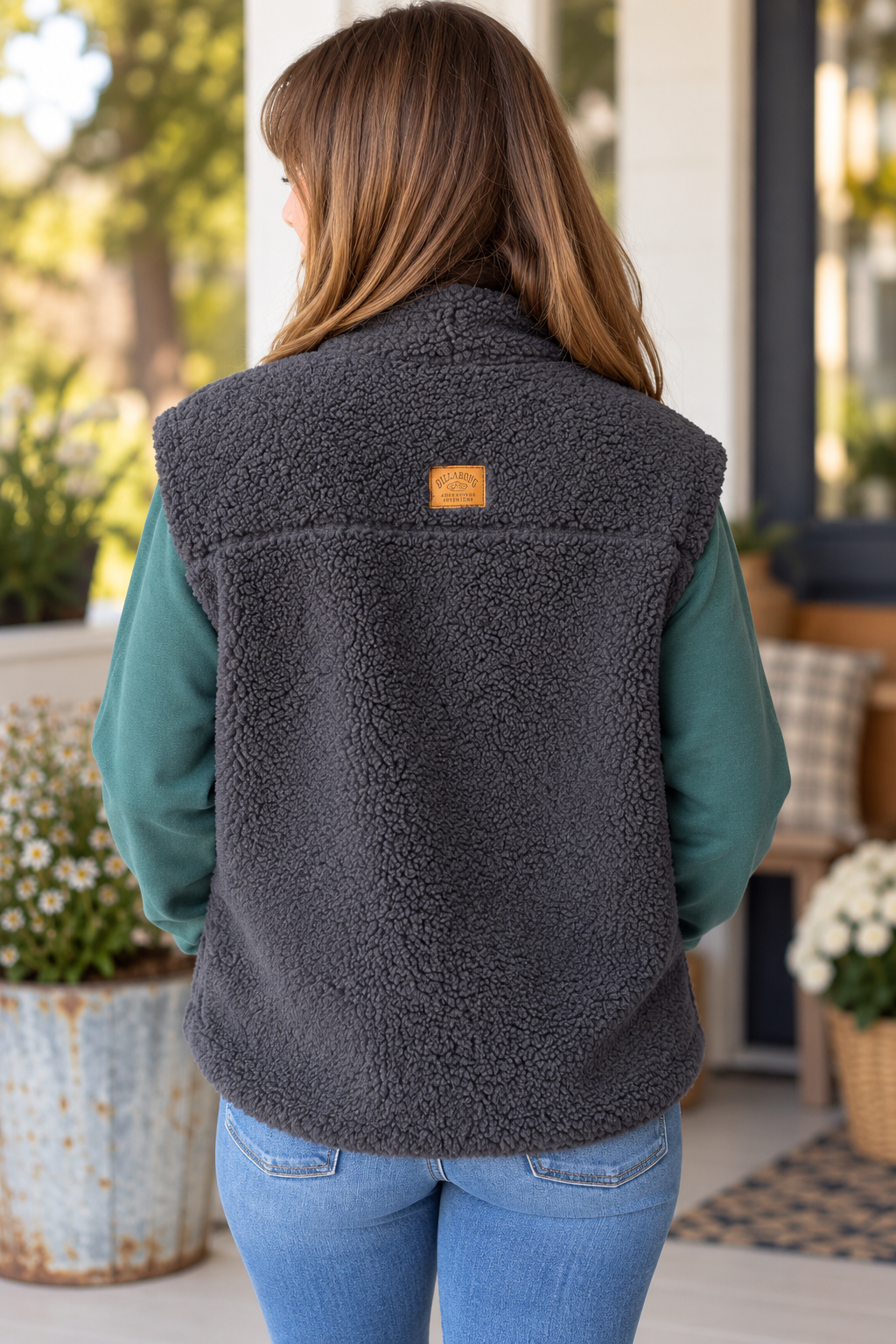 A woman viewed from behind wearing a dark gray sherpa fleece vest over a teal long-sleeve shirt and blue jeans, standing on a porch.