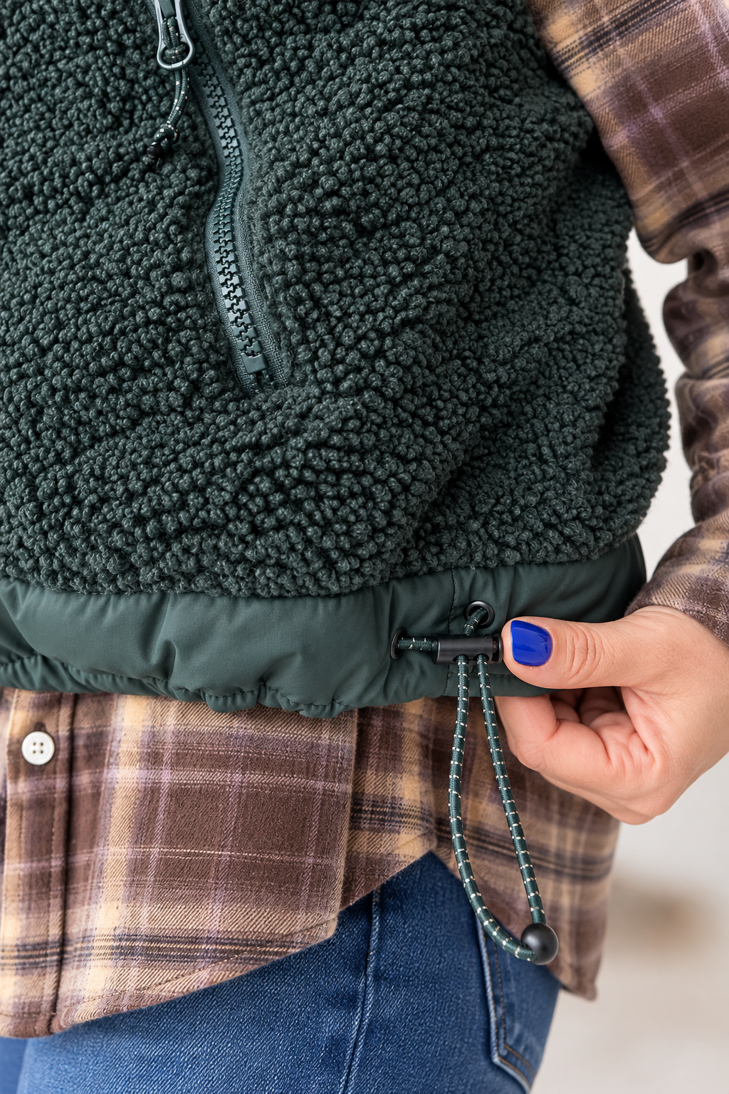 Close-up of a person wearing a dark green sherpa fleece vest over a brown plaid flannel shirt and blue jeans, with a hand adjusting the hem drawstring.