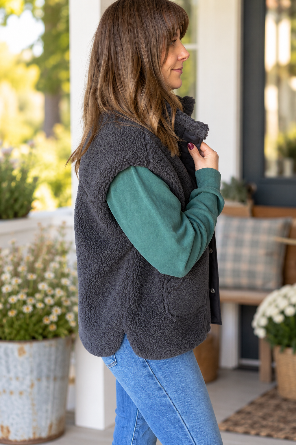 A woman wearing a dark gray sherpa fleece vest over a teal long-sleeve shirt and blue jeans, standing on a porch.