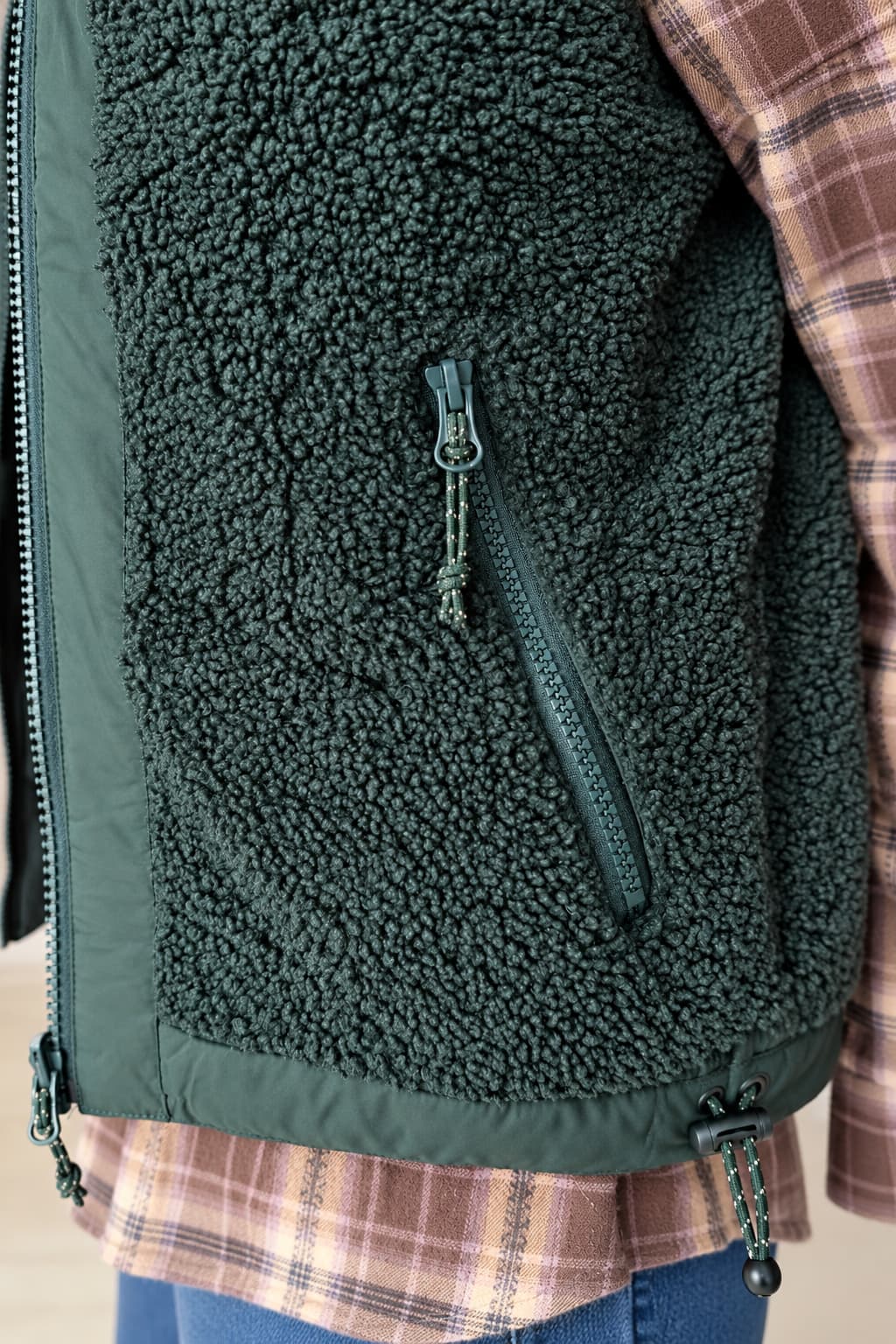 Close-up of a dark green fleece vest with a vertical zippered pocket, worn over a brown plaid shirt and blue jeans.