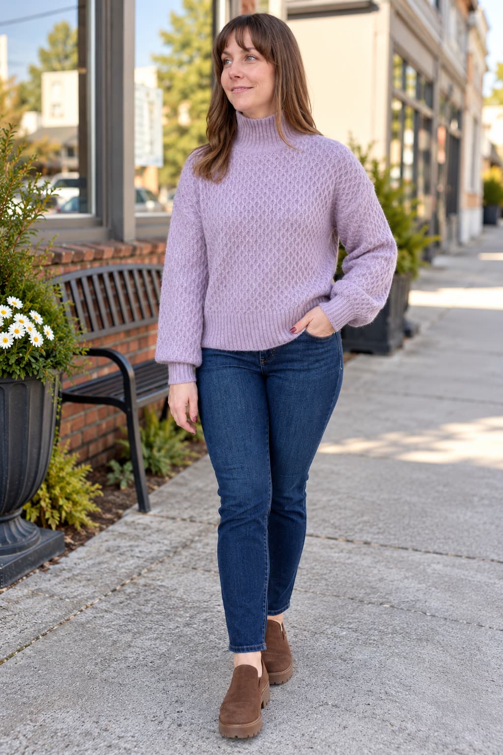 A woman wearing a lavender cable-knit mock neck sweater, dark blue skinny jeans, and brown suede boots, standing on a city sidewalk.