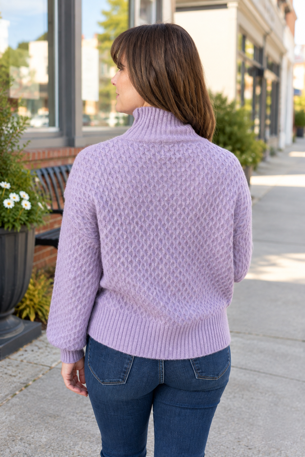 A woman viewed from behind wearing a lavender cable-knit turtleneck sweater and blue jeans, standing on a city sidewalk.