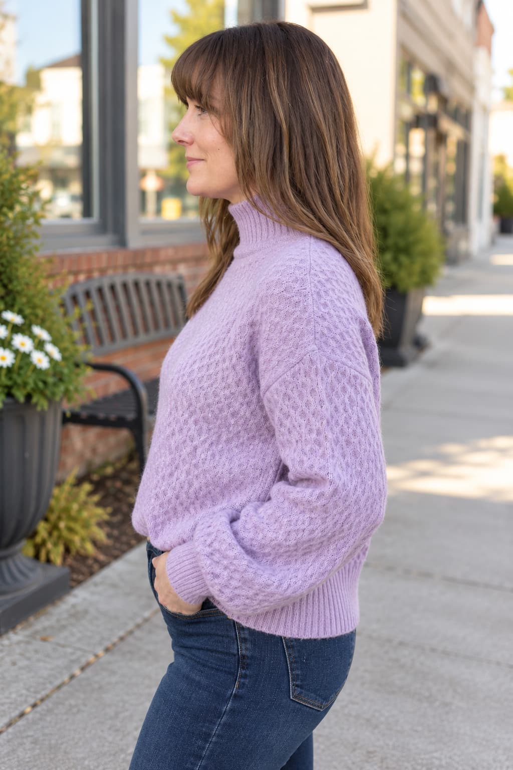 A woman wearing a lavender cable-knit mock neck sweater and dark blue jeans, standing on a city sidewalk.