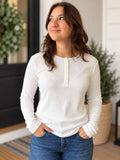 A woman wearing a white ribbed long-sleeve henley top with a button placket and blue jeans, standing in a home entryway.