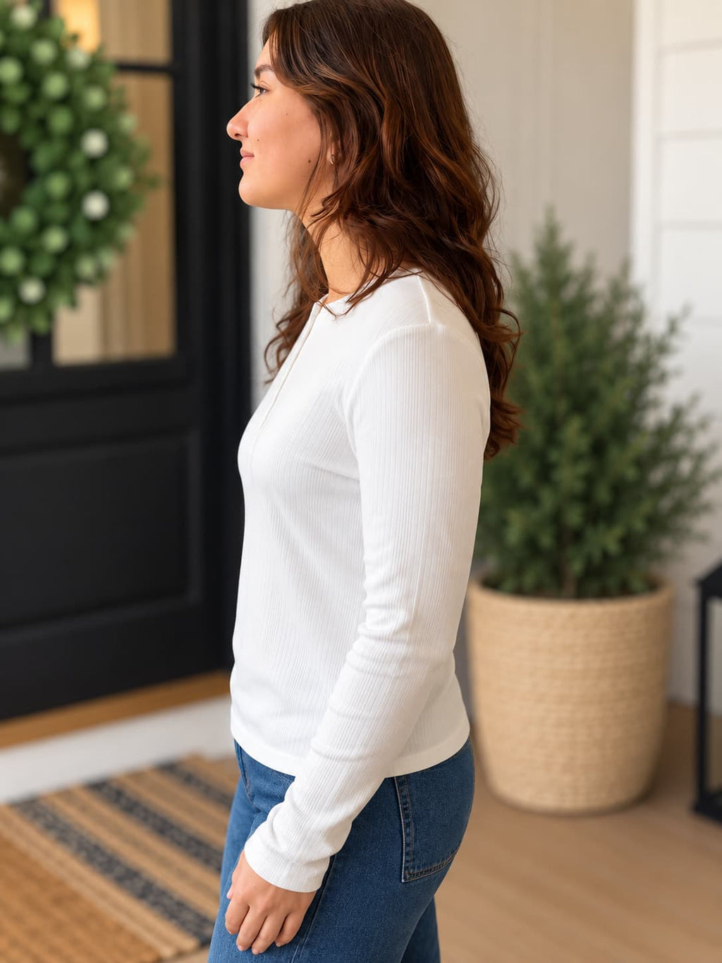 A woman in side profile wearing a white long-sleeve ribbed knit top and blue jeans, standing in a home entryway with a wreath and potted plants in the background.