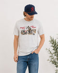 A man wearing a light gray graphic t-shirt, blue jeans, beige shoes, and a navy baseball cap with a red letter 'R' stands in a bright room with a bench, throw blanket, and potted plant.