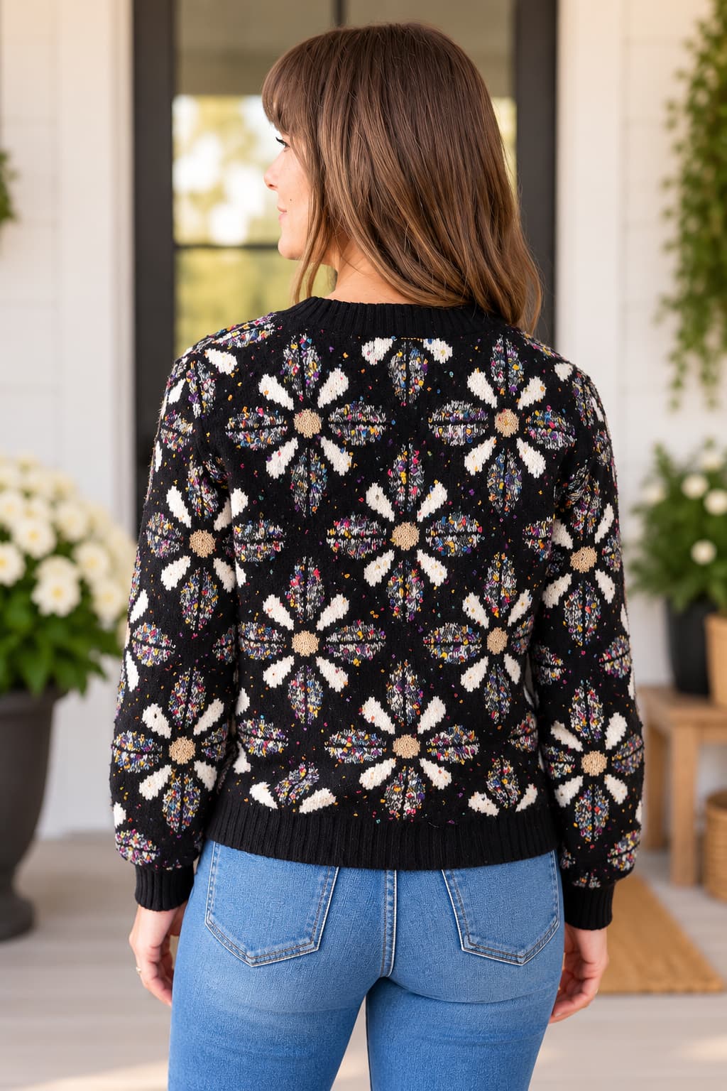 A woman viewed from the back wearing a black knit sweater with large cream and multicolor floral motifs, paired with blue jeans, in a bright porch setting.