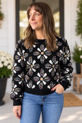 A woman wearing a black knit sweater with a large white and multicolor floral pattern, paired with blue jeans, standing on a porch.