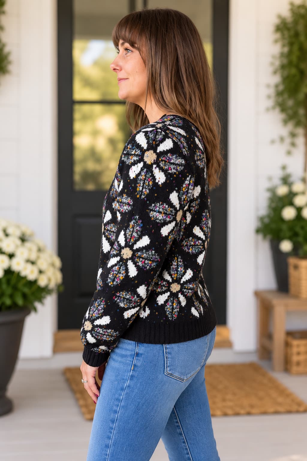 A woman wearing a black knit sweater with a large white and multicolor floral pattern, paired with blue jeans, standing on a porch.