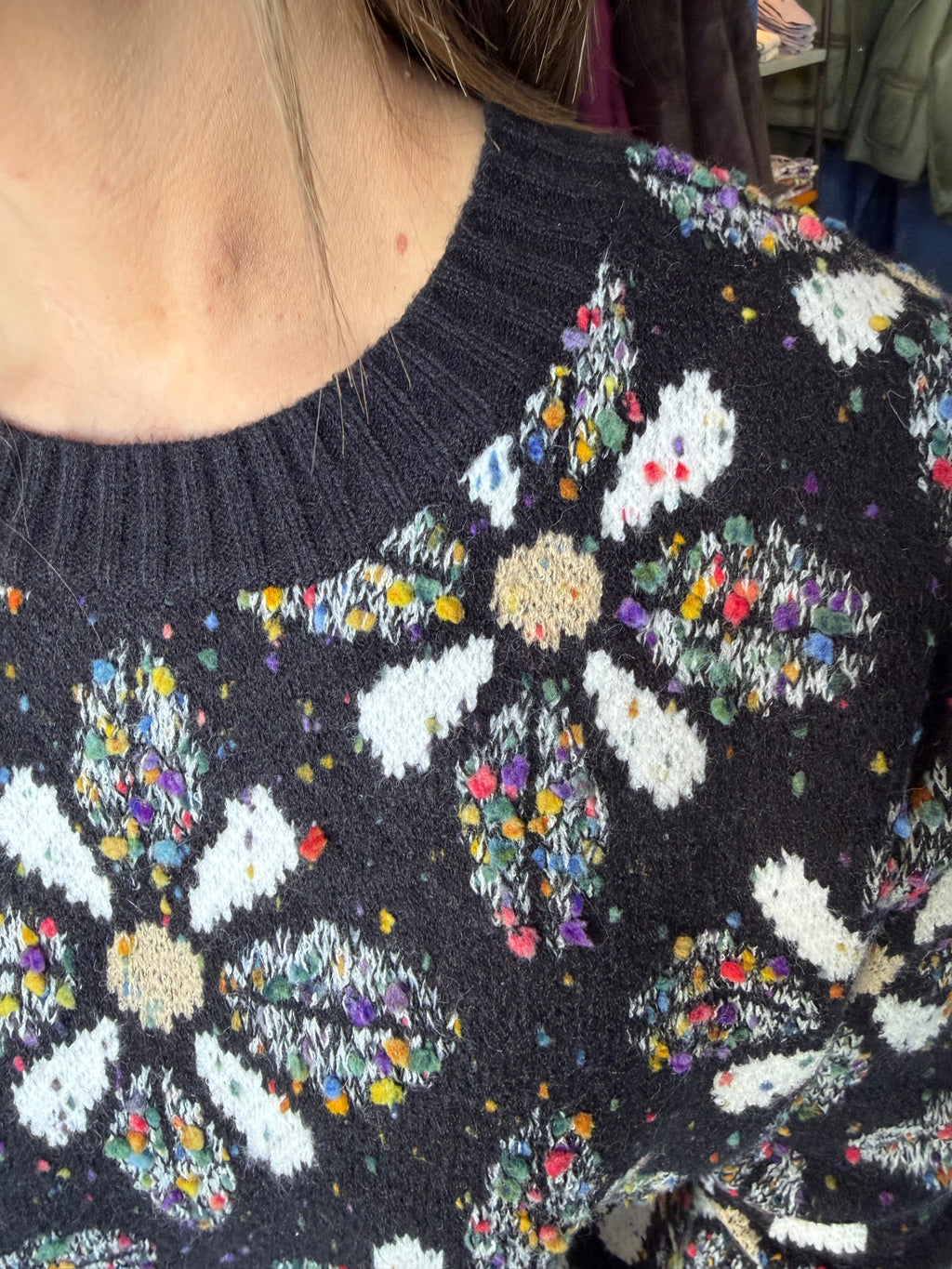 Close-up of a person wearing a dark charcoal knit sweater with large white floral motifs and dense multicolored yarn accents throughout.